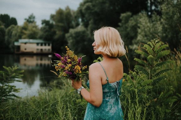 A beautiful blonde stands and holds a bouquet of freshly picked wildflowers in front of the lake