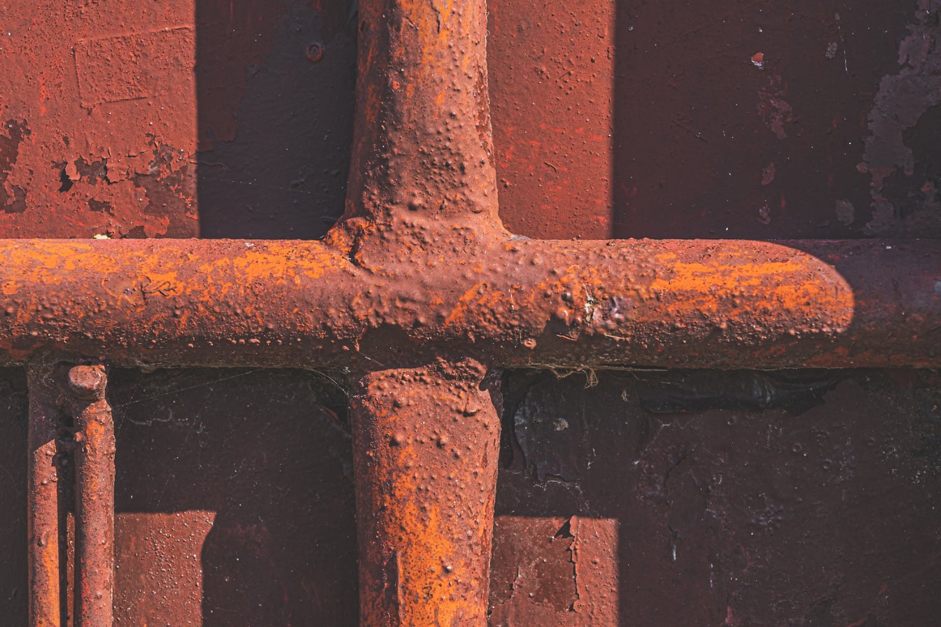 The texture of a rusty metal rod in semi-shade painted with primary reddish-brown paint that peels off