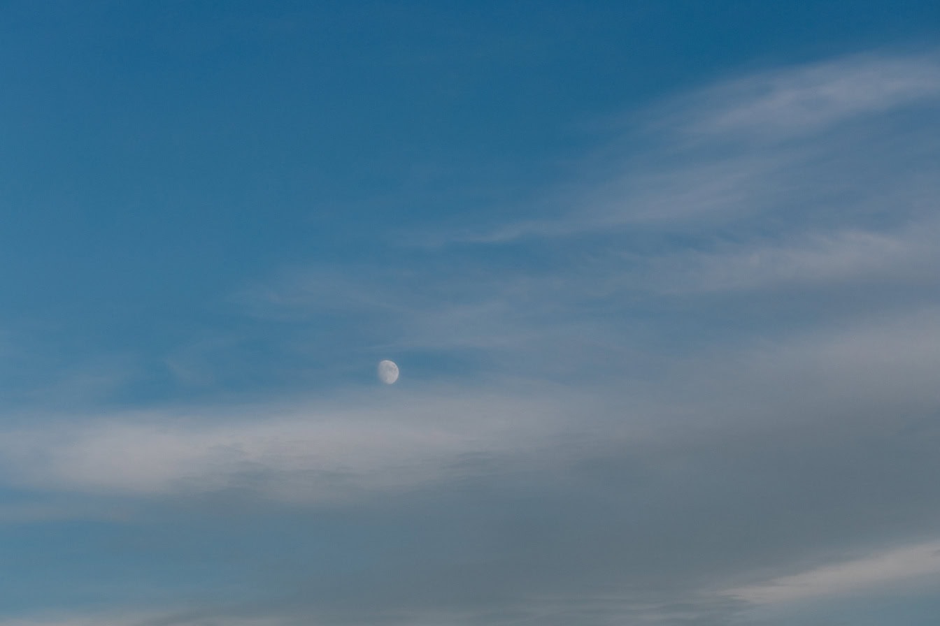 Blauwe hemel met mistige wolken en een kleine witte maan in de verte