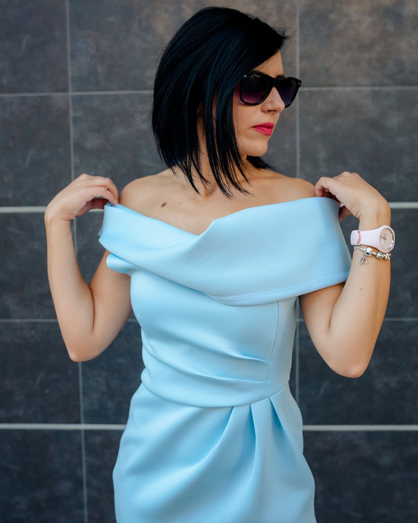 A woman wears sunglasses and poses with her hands on her shoulders while wearing an elegant blue dress