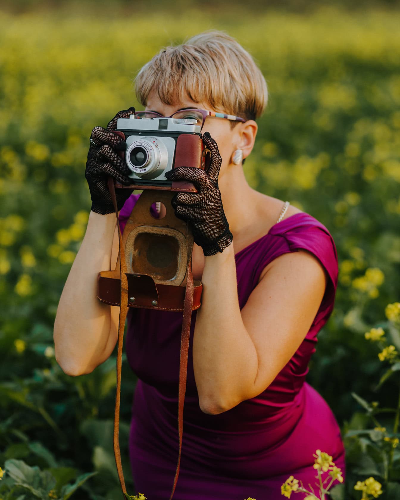 Femeie fotograf plină de farmec, cu coafură blondă scurtă în rochie mov, în timp ce face fotografie cu o cameră foto analogică