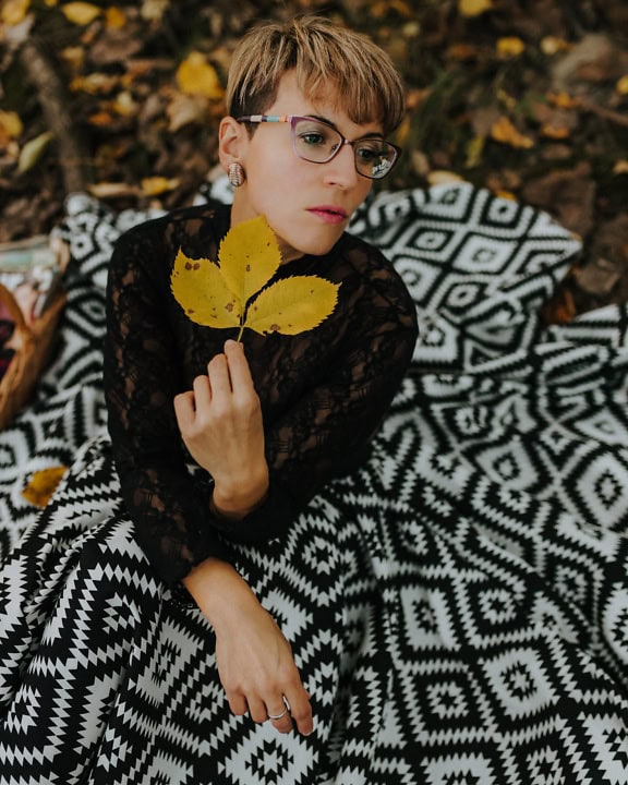 Бляскава дама с къса прическа, седнала в полупрозрачна елегантна дантелена риза и черно-бяла рокля и държаща жълтеникаво есенно листо