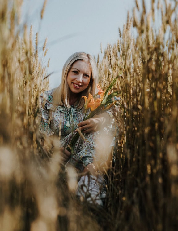Lächelnde junge Landfrau sitzt in einem Weizenfeld und hält einen Strauß orangefarbener Lilien in der Hand