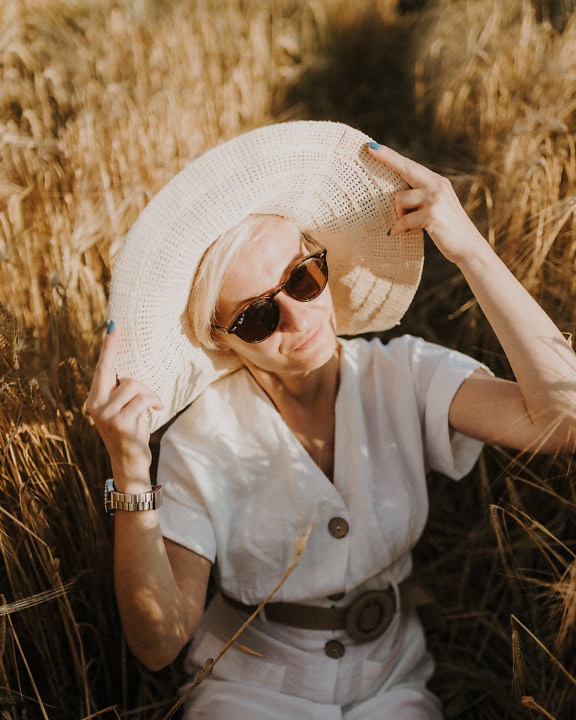 Блондинка с шапка и слънчеви очила позира, докато седи в житно поле в слънчев летен ден
