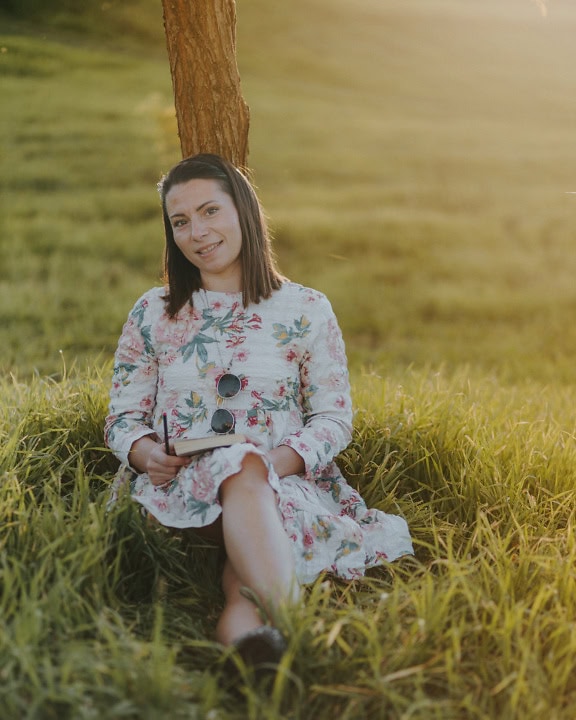 Porträt einer lächelnden jungen Frau, die im Gras sitzt und sich an einen Baum lehnt und ein Buch liest