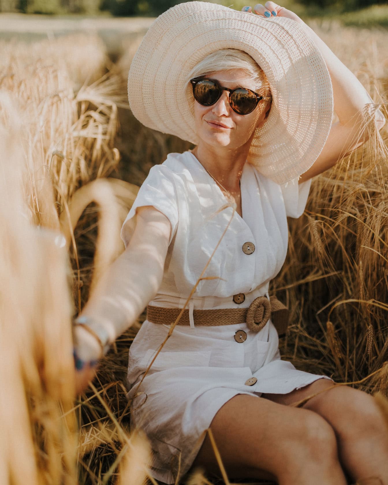 Wanita pirang glamor cantik dengan topi dan kacamata hitam berjemur di ladang gandum di akhir musim panas