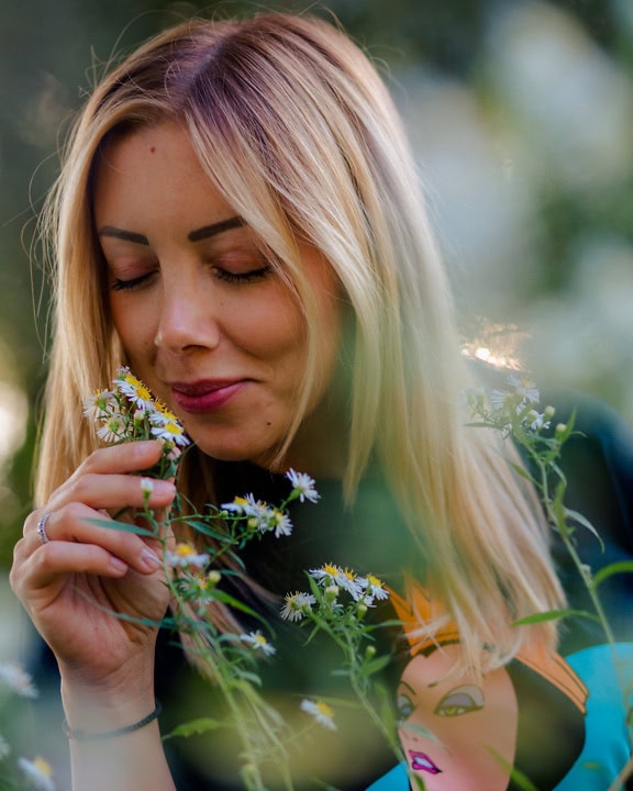一个年轻的金发女郎的肖像，有一张美丽的脸，同时在草地上闻着野生洋甘菊的花朵