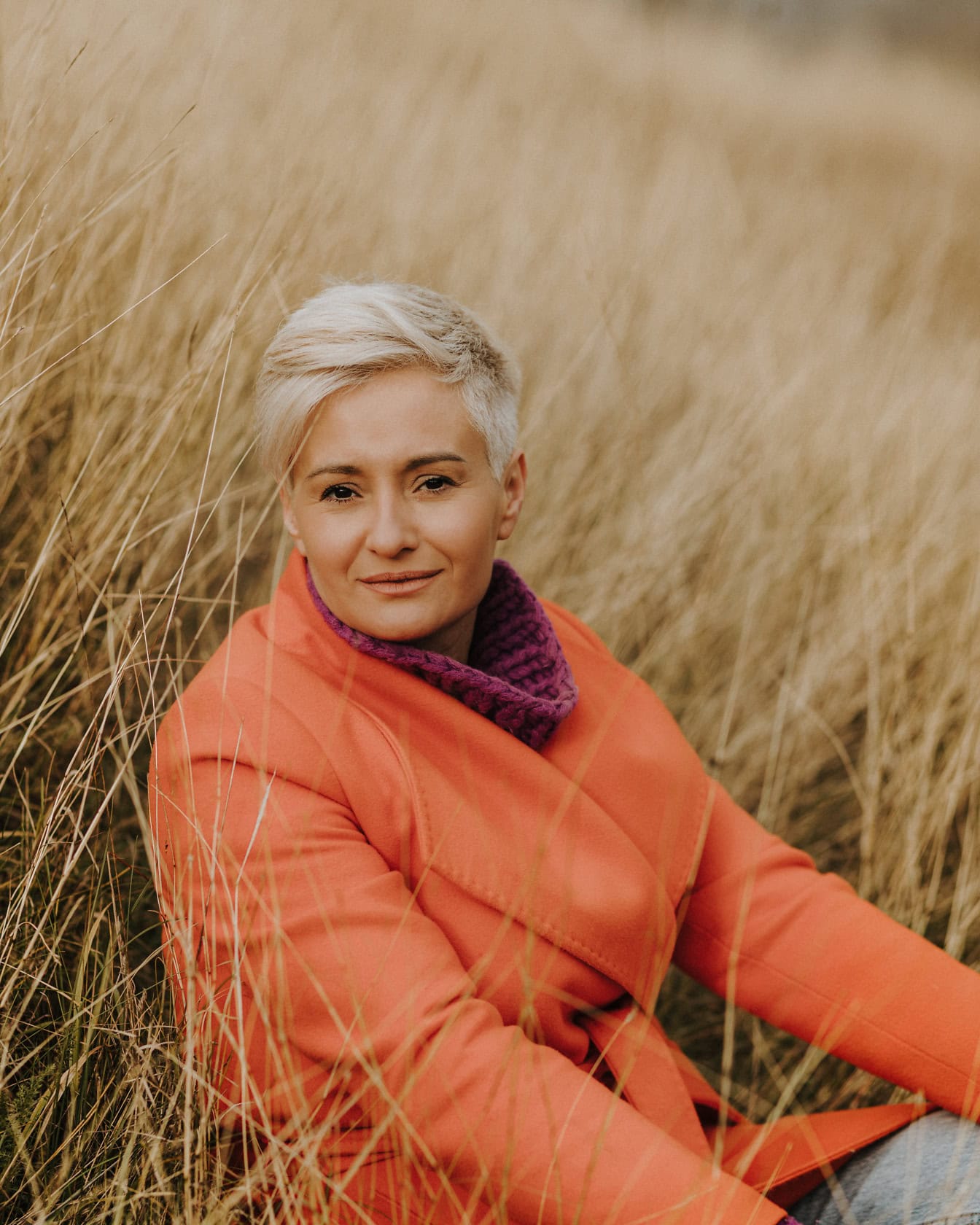 Portrait of a beautiful blonde with short haircut in an orange autumn coat while sitting in the dry tall grass