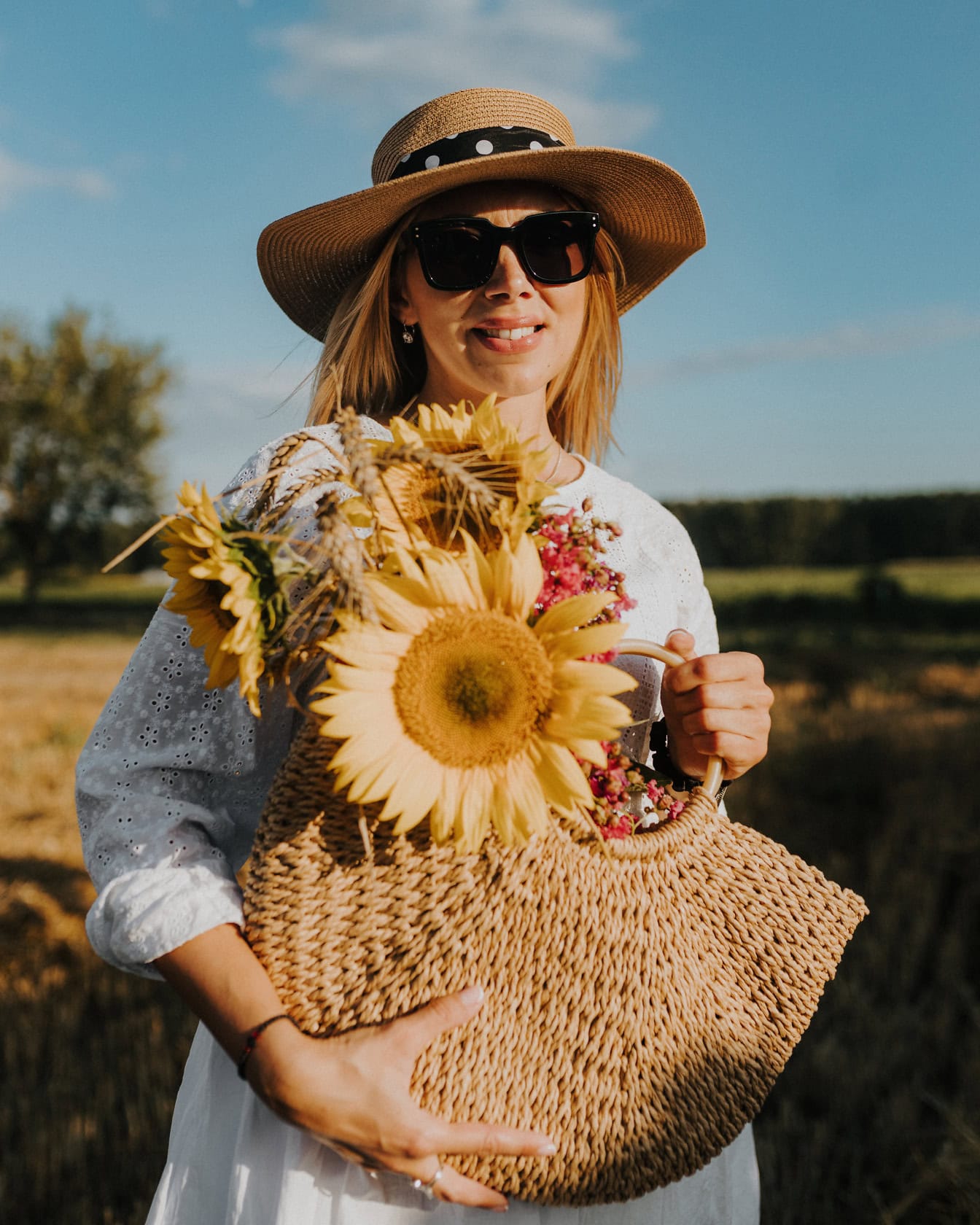 Красива млада блондинка носи сламена шапка и слънчеви очила, докато държи плетена слънчогледова чанта в полето