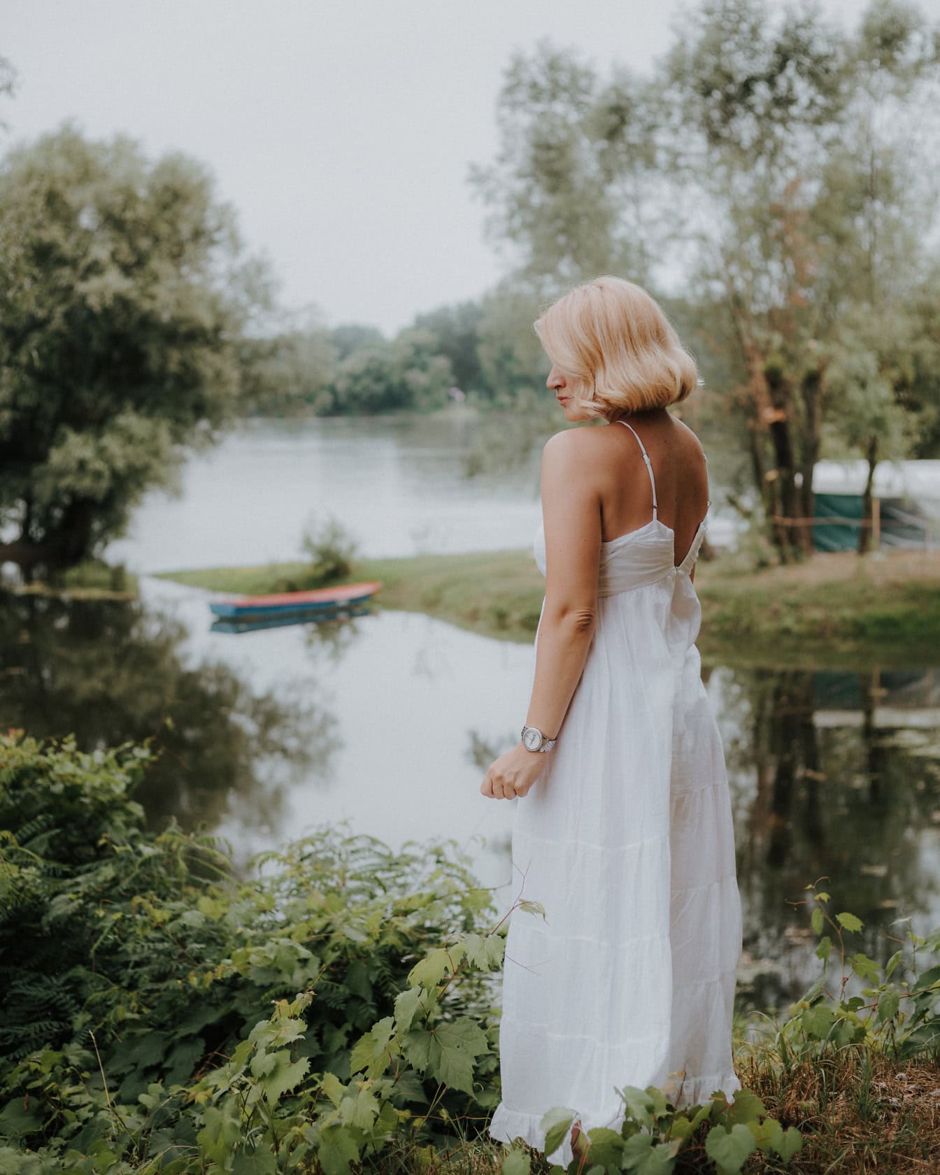 Seorang pengantin cantik dengan gaun pengantin gaya country berdiri di tepi danau