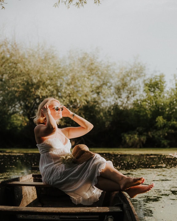 De mooie blonde zit in een houten boot in een witte kleding en zonnebaadt