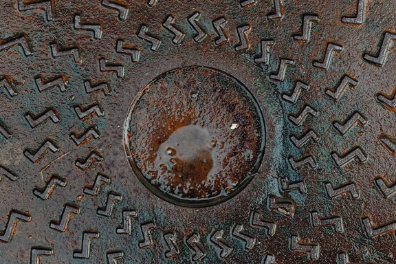 Текстурата на мокра повърхност на чугунена шахта с кръг в нея и украшение, напомнящо латинската буква z