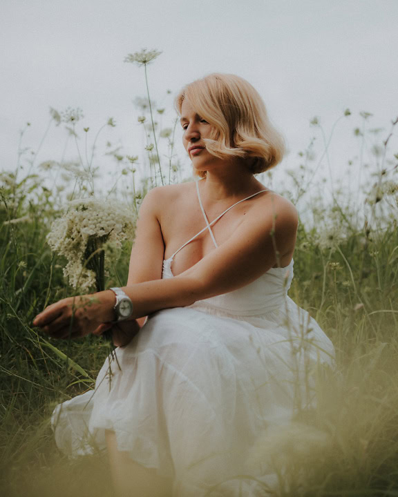 Приголомшливо красива молода блондинка в білій весільній сукні в стилі кантрі сидить на квітковому полі з букетом польових квітів в руці