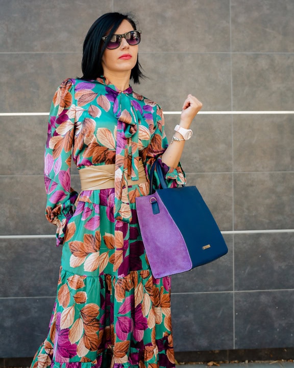 Porträtt av en affärskvinna som bär en elegant färgrik klänning och solglasögon medan hon rymmer en lila-blå läderhandväska