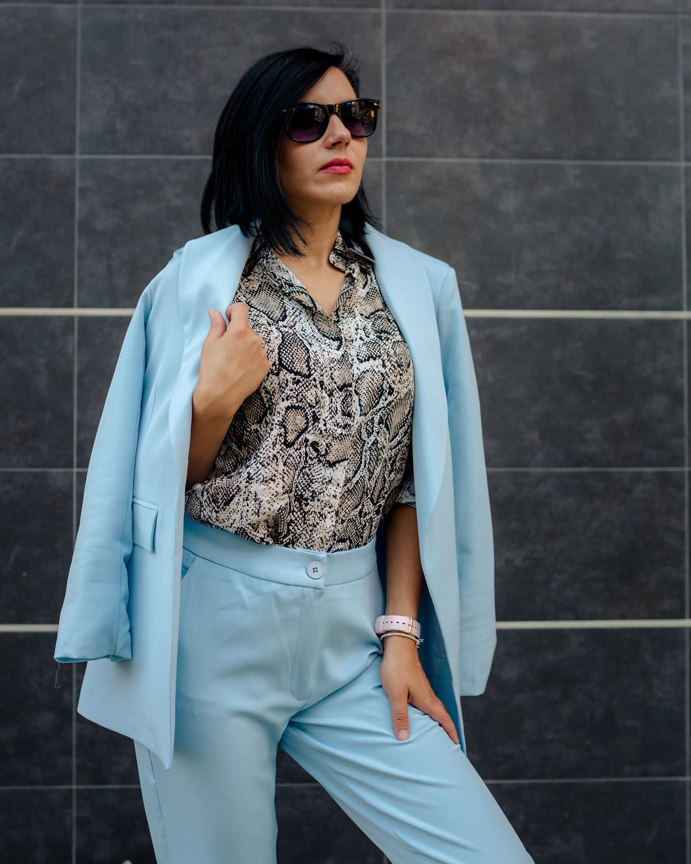 A businesswoman poses in sunglasses and elegant light blue pants and a coat and a shirt with a snakeskin pattern
