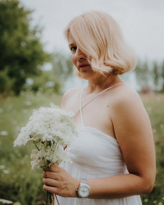 Smuk blond kvinde i hvid kjole på eng med en buket hvide vilde blomster