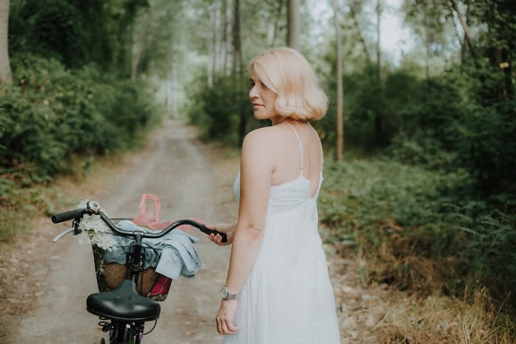 Vakker blond kvinne i hvit kjole med sykkel på skogsstien
