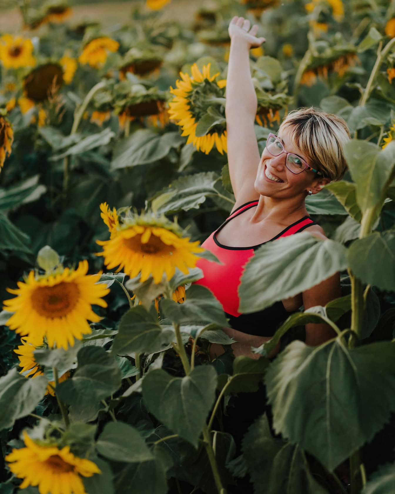Porträt einer glücklich lächelnden jungen Frau mit kurzer blonder Frisur in einem Sonnenblumenfeld mit erhobener Hand in der Luft