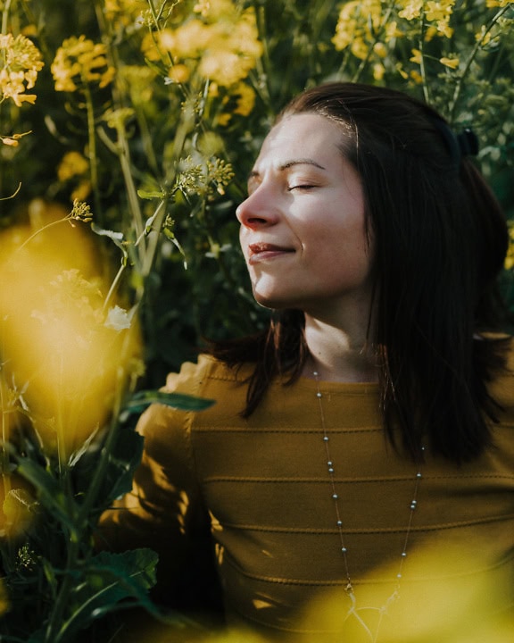 Porträtt av en förtjusande lycklig brunett i en gul tröja som ler i ett fält av gulaktiga blommor medan hon luktar på det