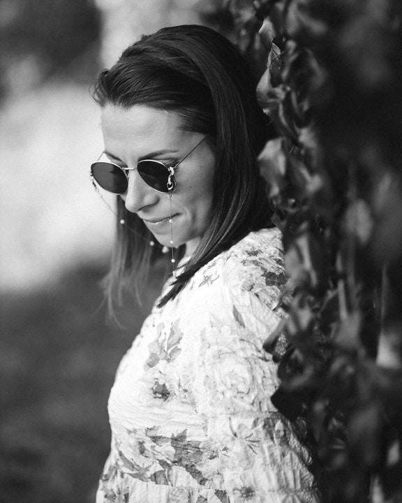 Професионална черно-бяла портретна снимка на млада жена, носеща слънчеви очила в стил Джон Ленън, докато се обляга на бръшлянов храст
