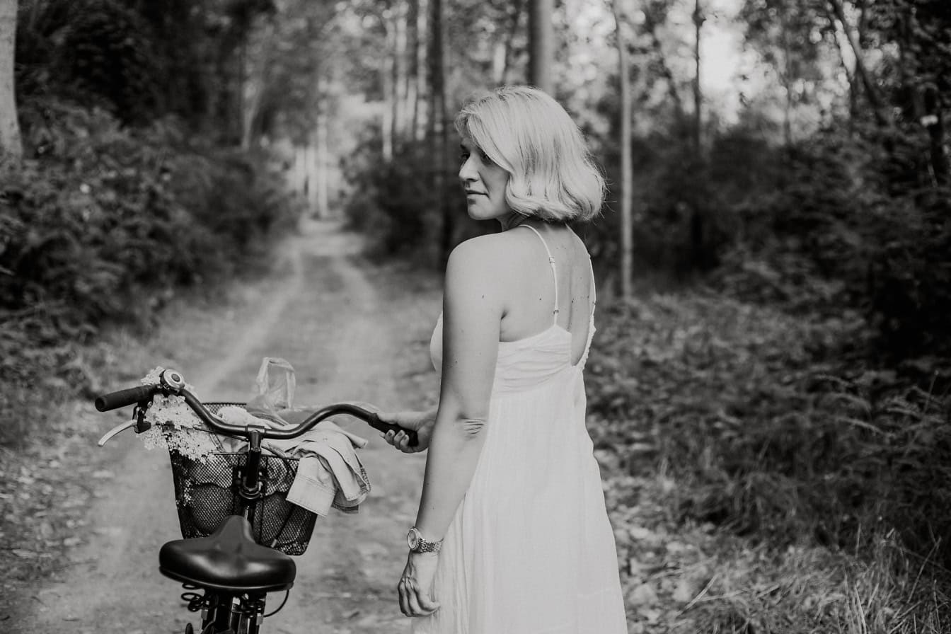 Professzionális fekete-fehér fénykép egy gyönyörű nőről fehér ruhában, kerékpárral az erdőben