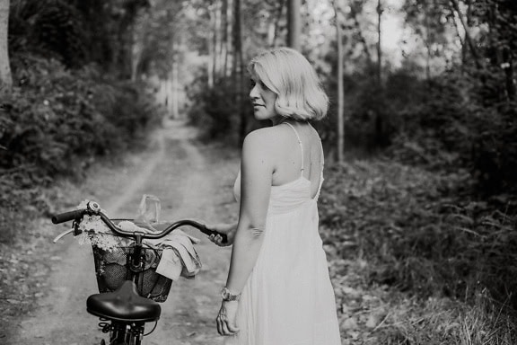 Professional black and white photograph of a beautiful woman in a white dress with a bicycle in the woods