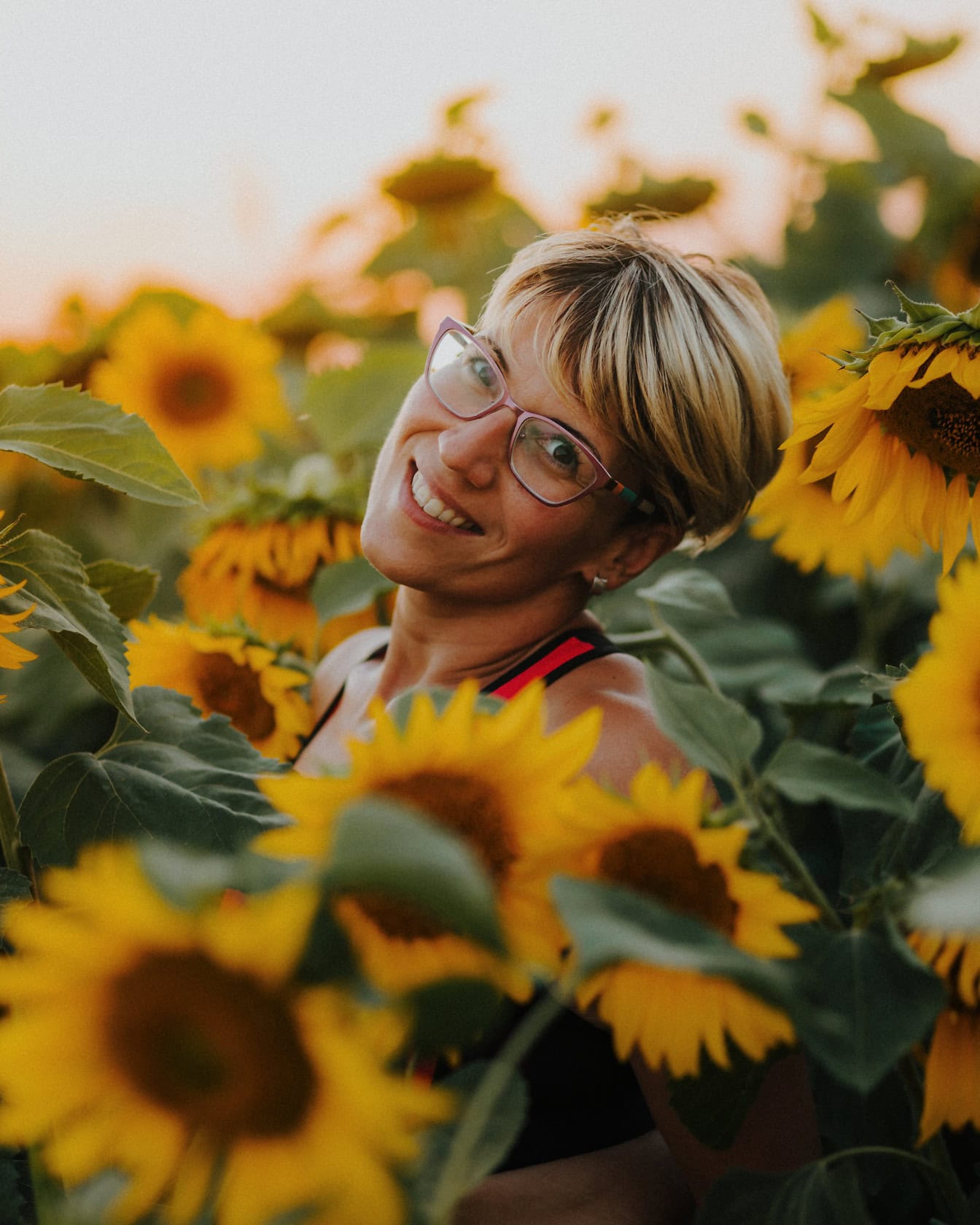 Professionellt porträtt av en lycklig kvinna med kort blond frisyr som står i ett fält av solrosor
