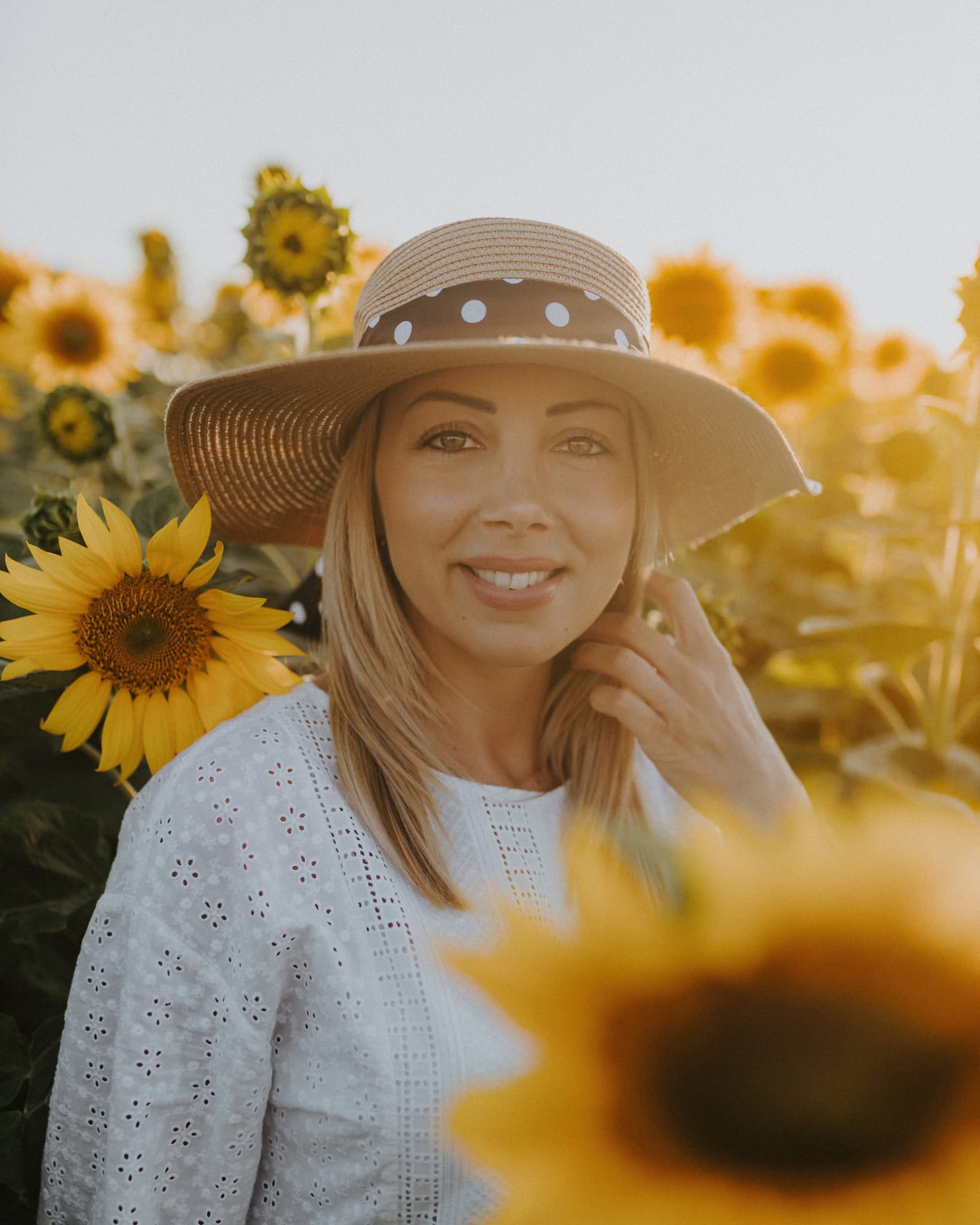 Fotografie portret profesională cu o tânără frumoasă, fotomodel, purtând o pălărie de paie într-un câmp de floarea-soarelui, cu razele soarelui ca lumină de fundal
