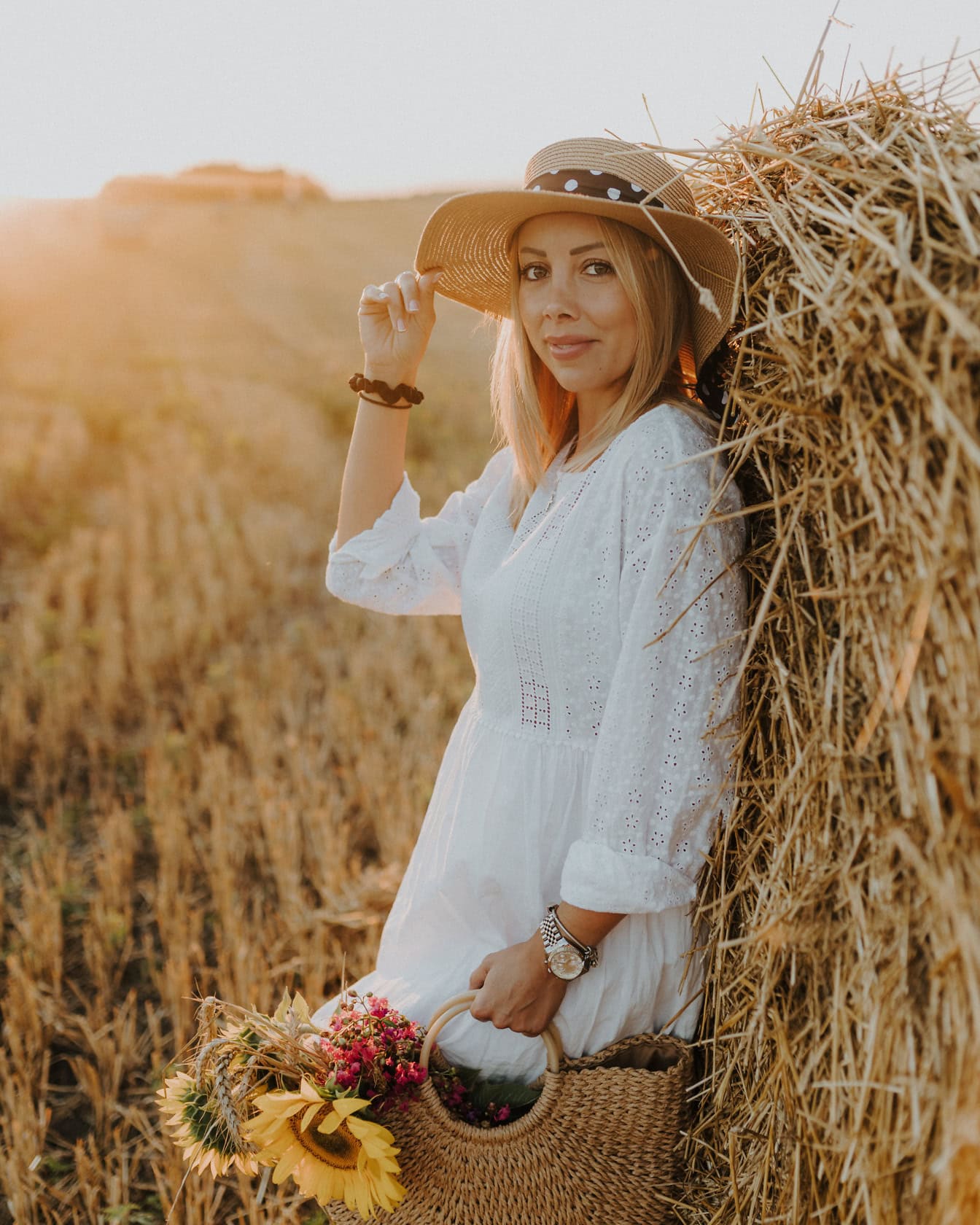 Portrait of a cowgirl in a country-style white dress and straw hat while she leaning against a haystack in a sunny field