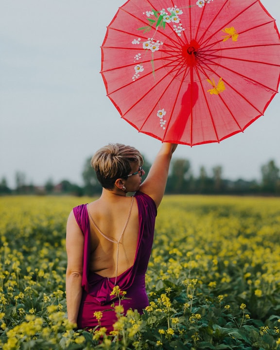 一位穿着紫色连衣裙的开朗迷人的女士的肖像，她站在花田中，高举着一把红色的雨伞