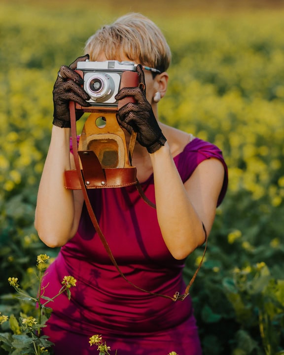 Fotografer pirang berpakaian glamor memegang kamera foto analog dengan kedua tangan dengan sarung tangan renda di tangannya