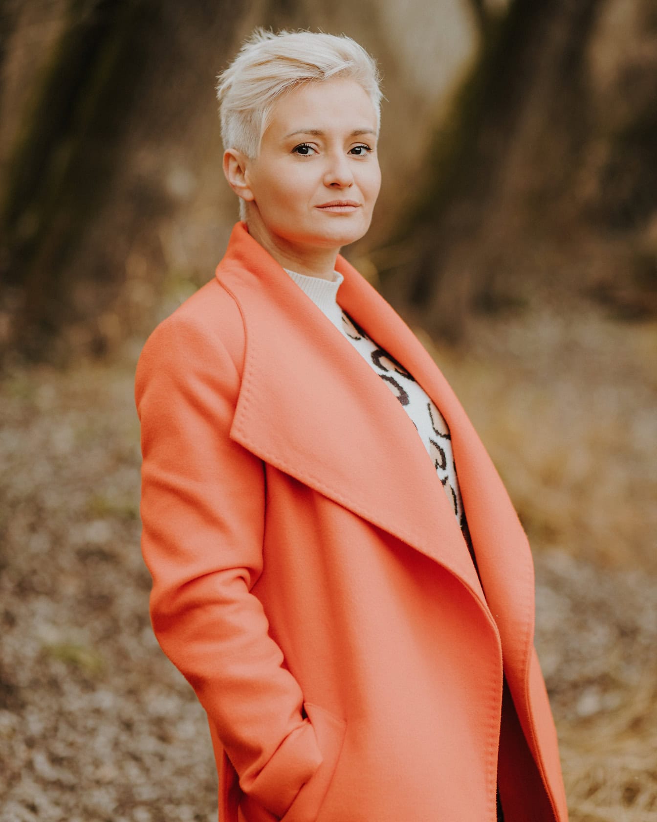 Beautiful blonde woman with short hair in an orange coat poses for a picture while standing