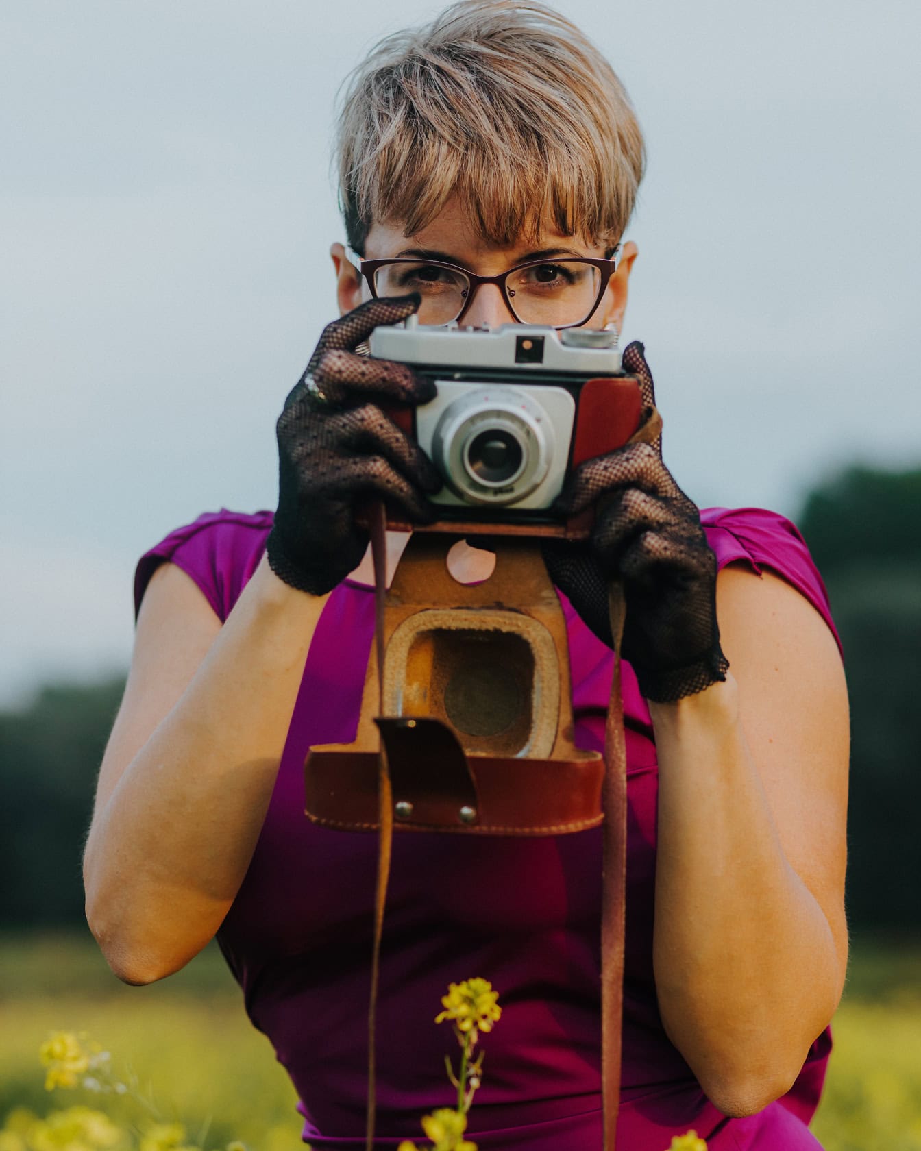 Nữ nhiếp ảnh gia ăn mặc quyến rũ trong chiếc váy màu tím cầm một chiếc máy ảnh analog cũ với găng tay ren và chụp ảnh