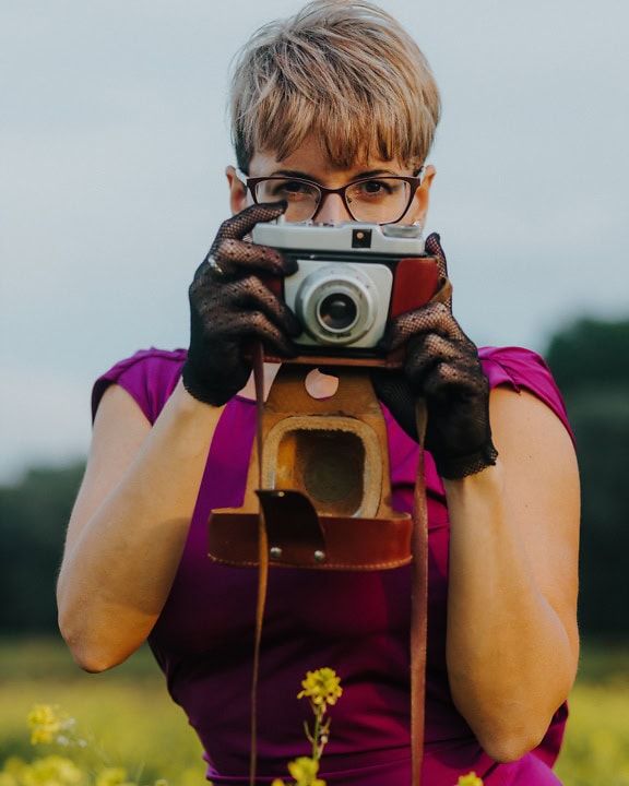 Okouzlující fotografka ve fialových šatech, držící starý analogový fotoaparát s krajkovými rukavicemi a fotografující
