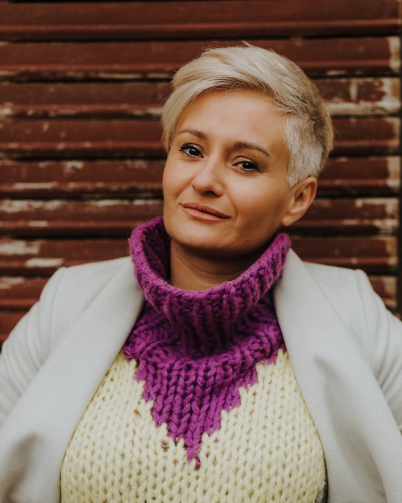 Portrait of a confidently smiling blonde woman with short hair wearing a purplish handwoven sweater and a white jacket