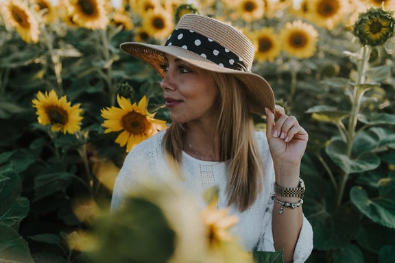 Porträtt av en underbar ung kvinna på landet som bär en hatt och vit klänning i ett fält av solrosor