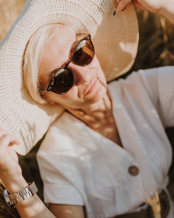 Obličejový portrét glamour lady v béžovém klobouku, bílých šatech a slunečních brýlích za slunečného letního dne