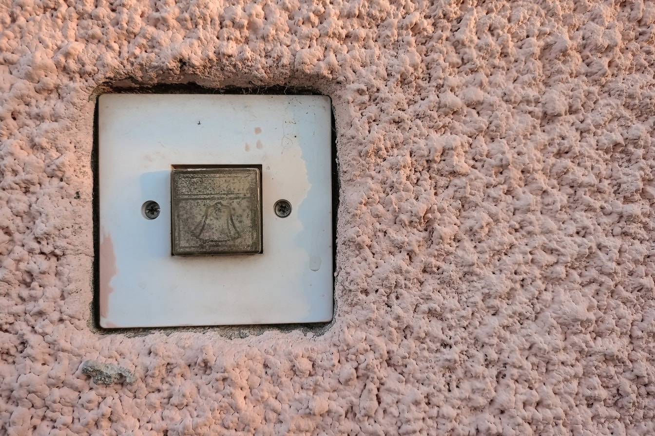 Zbliżenie przycisku dzwonka na białym plastikowym przełączniku na szorstkiej różowawej ścianie cementowej