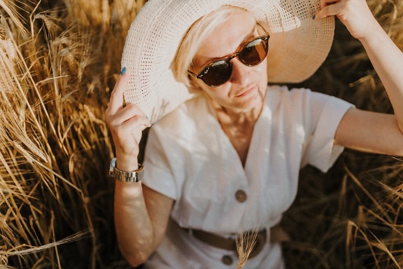 A high-angle portrait of a beautiful woman with short blonde hair with a glamorous white hat and dress and sunglasses