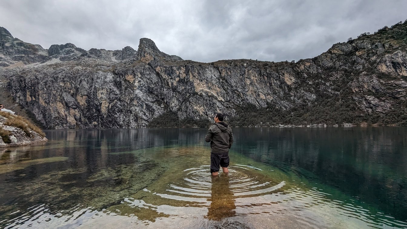 Photograph of a man standing in Charup lake in natural park in Peru with a majestic landscape of mountains in the background