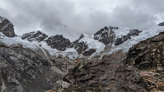 Homem à distância sentado no penhasco da montanha com picos nevados na montanha no fundo no parque natural do Peru