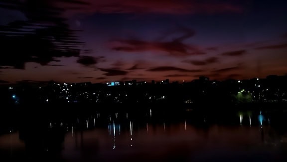 Stadshorisont på natten med en reflektion av stadsljus på ett lugnt vatten med mörk purpurröd himmel som bakgrund