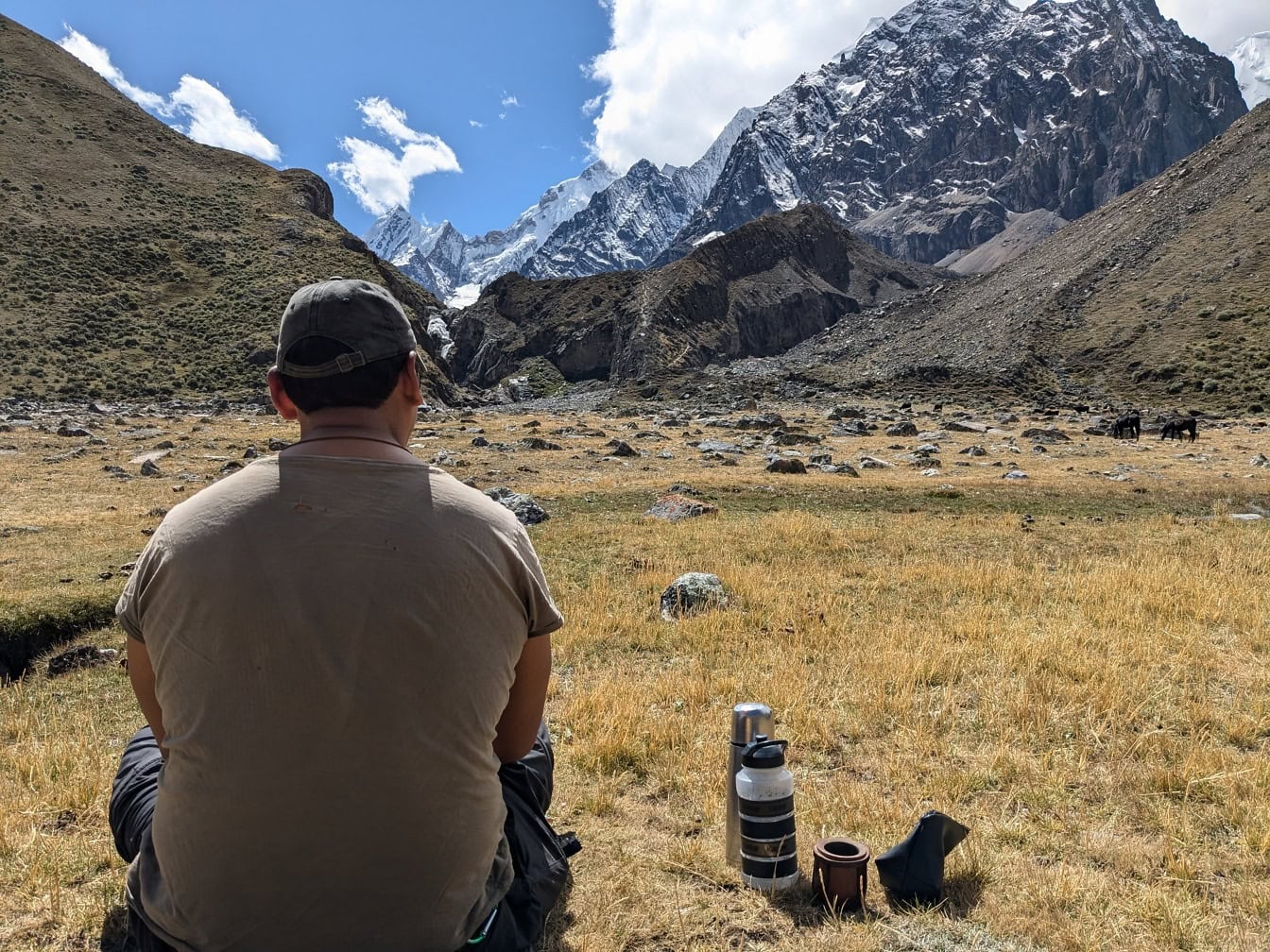Mies istumassa pellolla, jonka taustalla on vuoria, Cordillera Huayhuashin vuorijonossa Perussa;