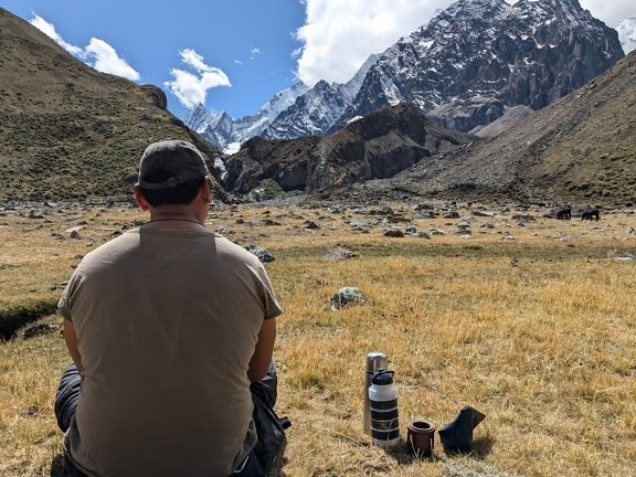 Pria duduk di lapangan dengan pegunungan di latar belakang di pegunungan Cordillera Huayhuash di Peru