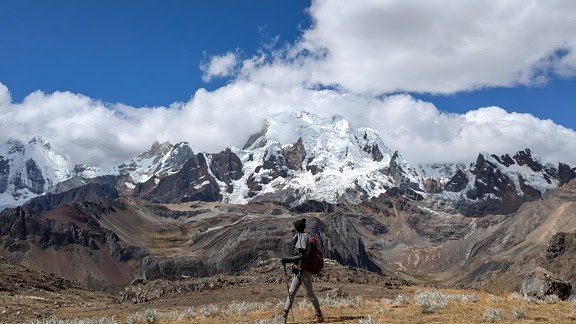 Hiker walking in a field with a snowy mountain peaks of a a Cordillera Huayhuash mountain range in the Andes of Peru in the background