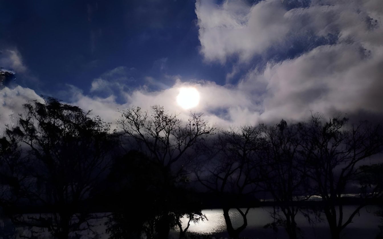 Sol skinner gennem skyer over vand med silhuet af træer på land i skumringen