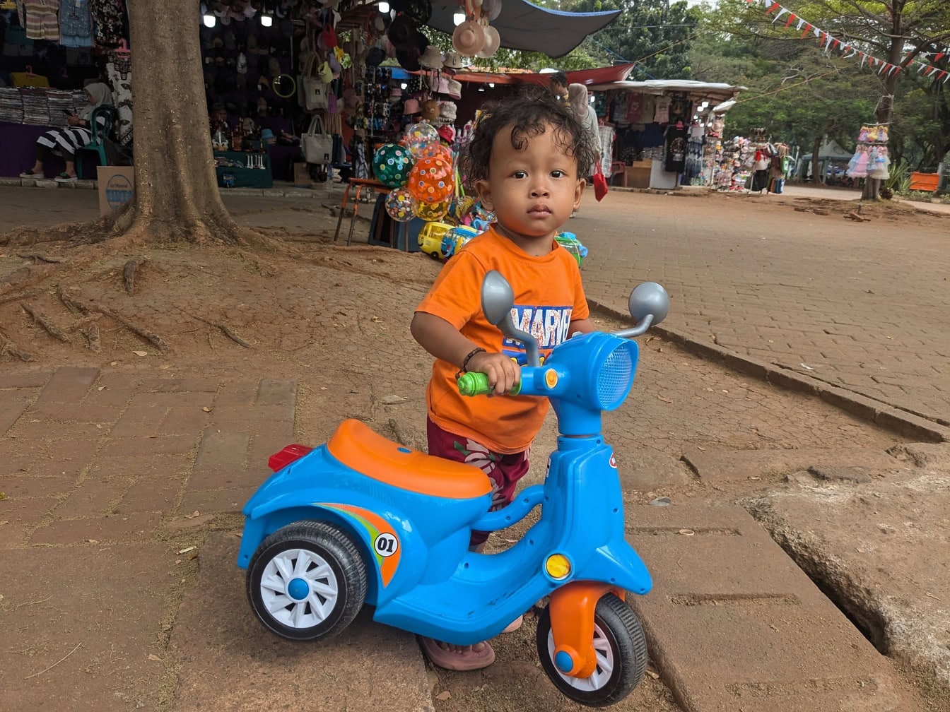 Seorang anak balita yang lucu berdiri di samping sepeda motor mainan biru di jalan Jakarta di Indonesia