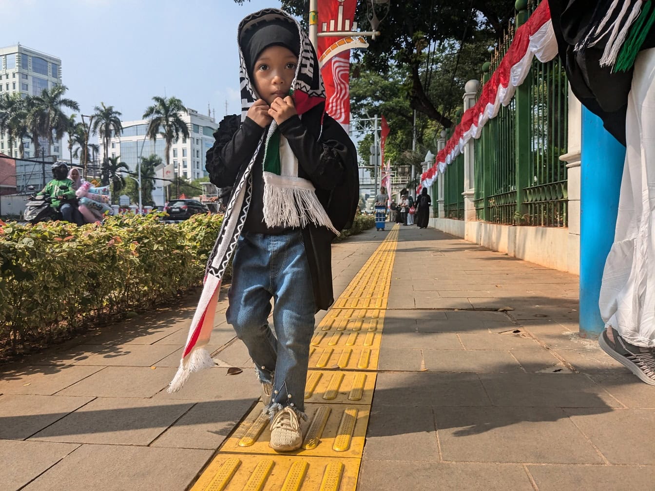 A young Indonesian boy with a sports fan scarf on his head walks on the sidewalk