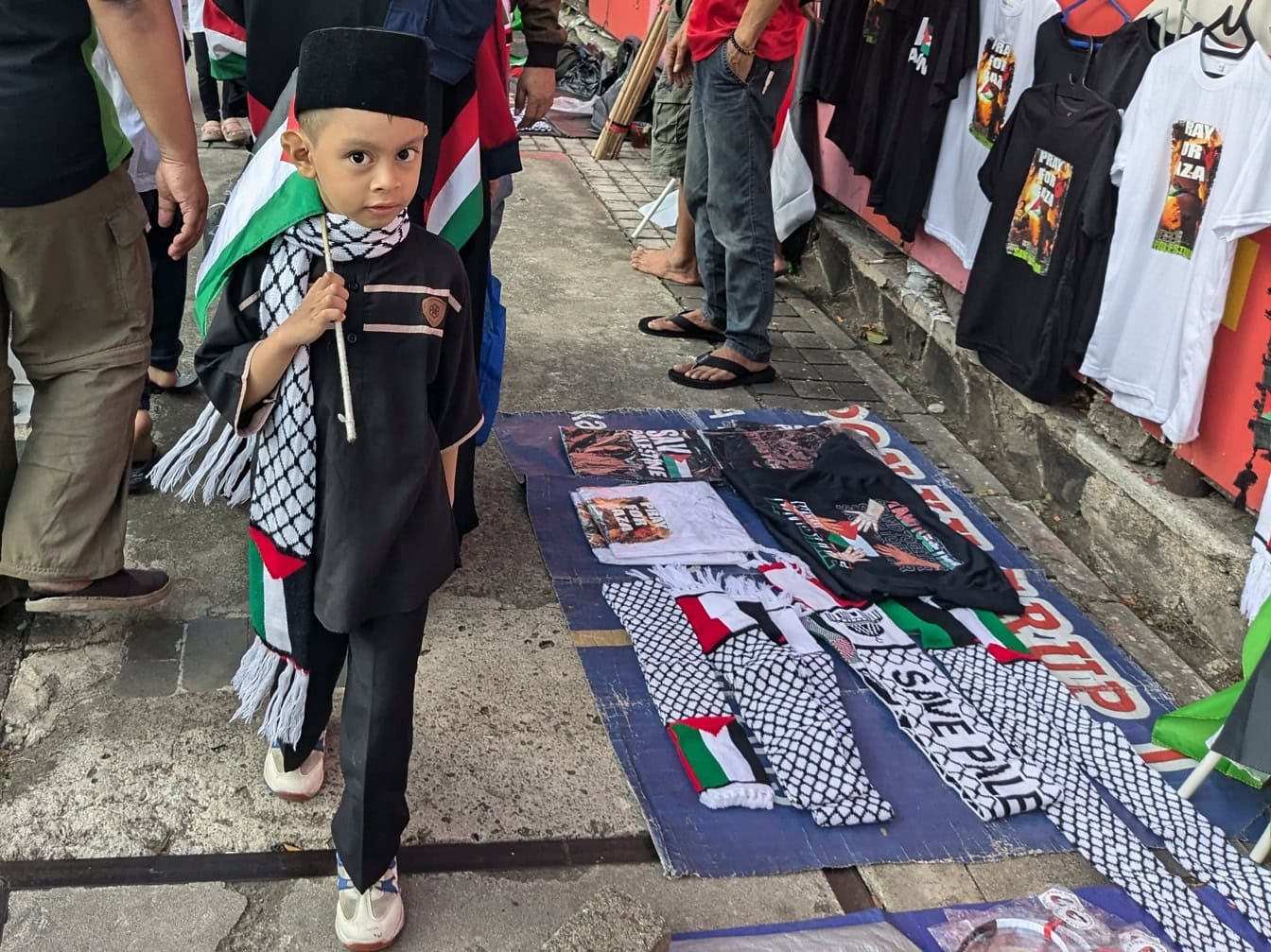 Seorang anak laki-laki berbendera Indonesia berdiri di samping T-shirt yang dijual di pasar jalanan