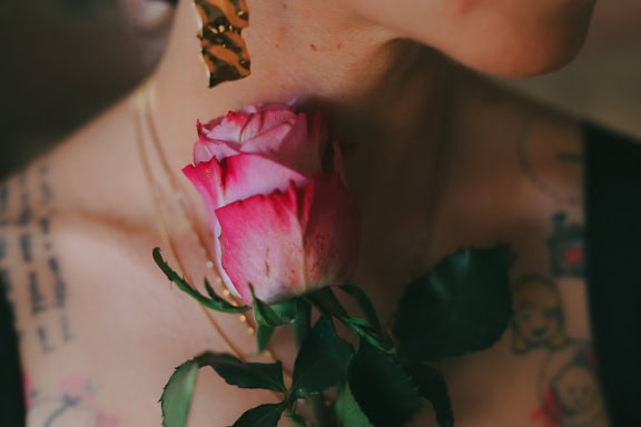 Close-up of a a pinkish rose bud next to a neck of woman with tattoos